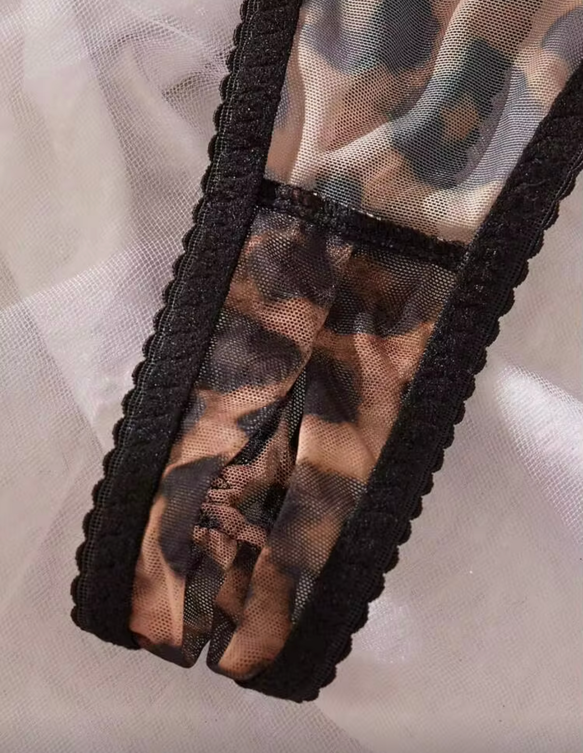 A close-up of a sheer crotchless lingerie underwear strap with a brown and black leopard print pattern, finished with black scalloped trim, shown against a white fabric background.
