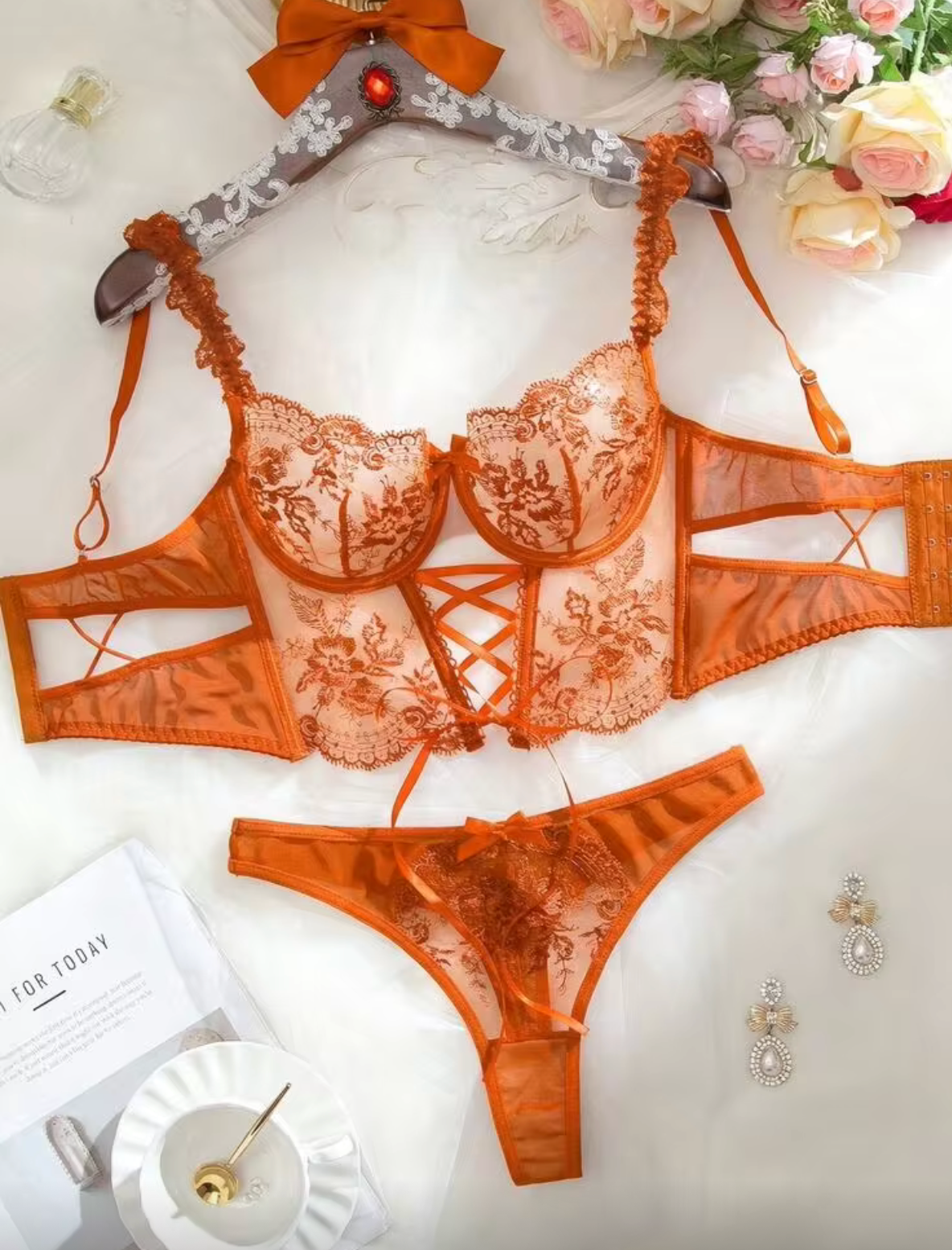An orange lace lingerie set consisting of a sheer underwire bra and matching panties, displayed on a white fabric background with decorative flowers and a hanger.