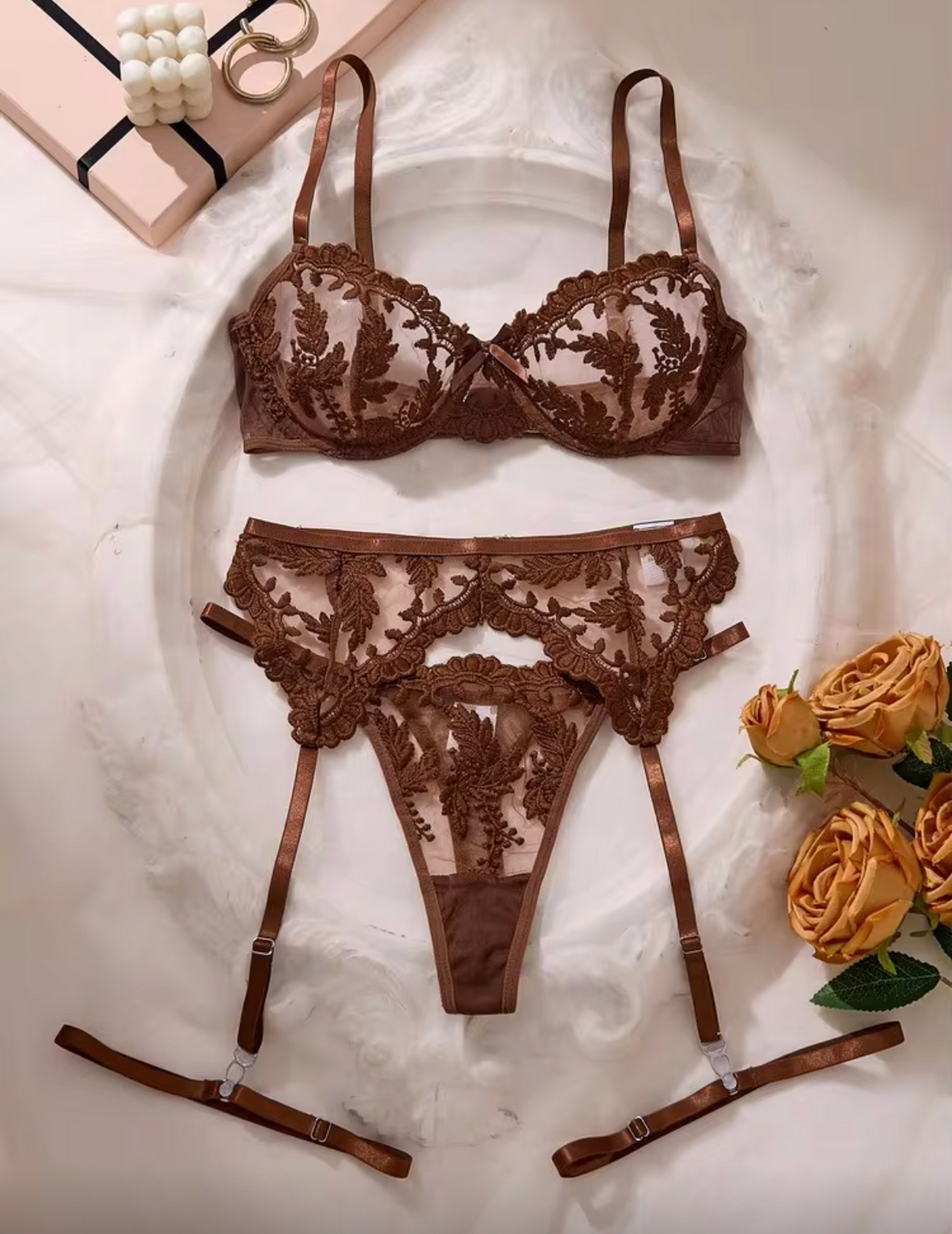 A three-piece brown lace lingerie set laid flat on a white fabric background, featuring a bra, matching panties, and garter straps. Decorative gold rings and yellow roses are placed around the set.