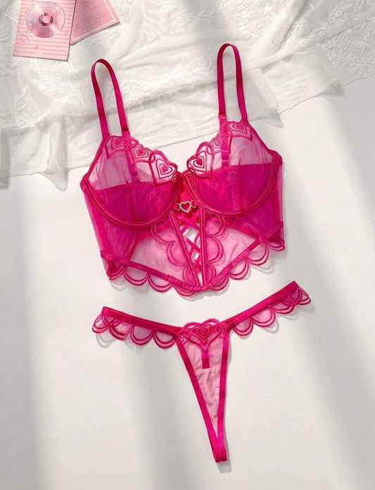 A two-piece lingerie set in vibrant hot pink, featuring a sheer mesh and lace bralette with scalloped edges and a matching thong, laid flat on a white textured fabric background.