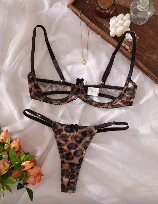 A leopard-print lingerie set consisting of a bralette and matching thong, laid flat on white fabric with decorative props including flowers, a wooden tray, and a candle. The set is labeled as 'Brown' in the color selector below.