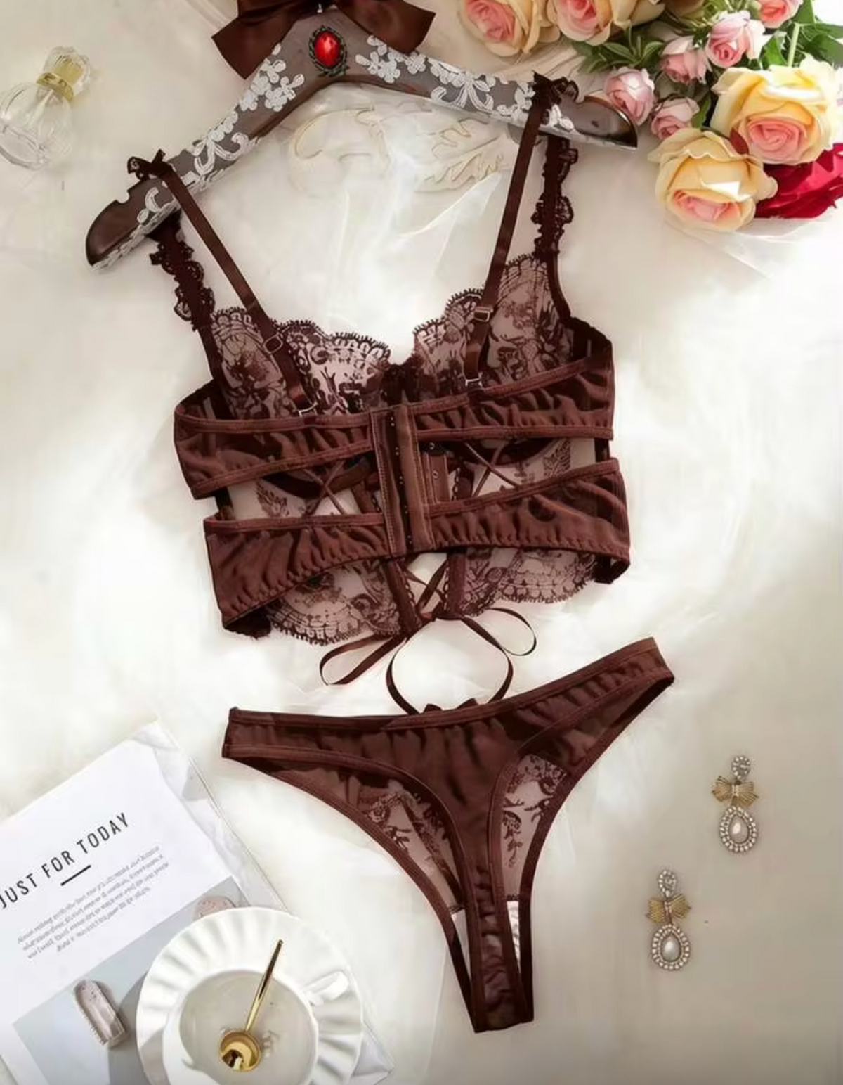 A two-piece brown lace lingerie set consisting of a bralette and matching panty, laid flat on a white fabric background with decorative flowers, jewelry, and a cup nearby.