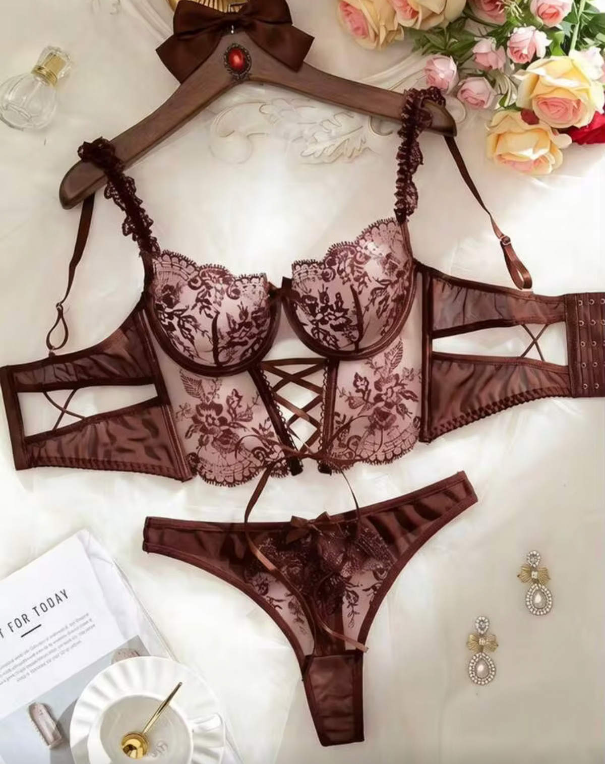 A three-piece lace lingerie set in brown featuring a lace bra, matching panties, and a lace belt with a bow, displayed on a hanger over a white fabric background with decorative flowers and a teacup.