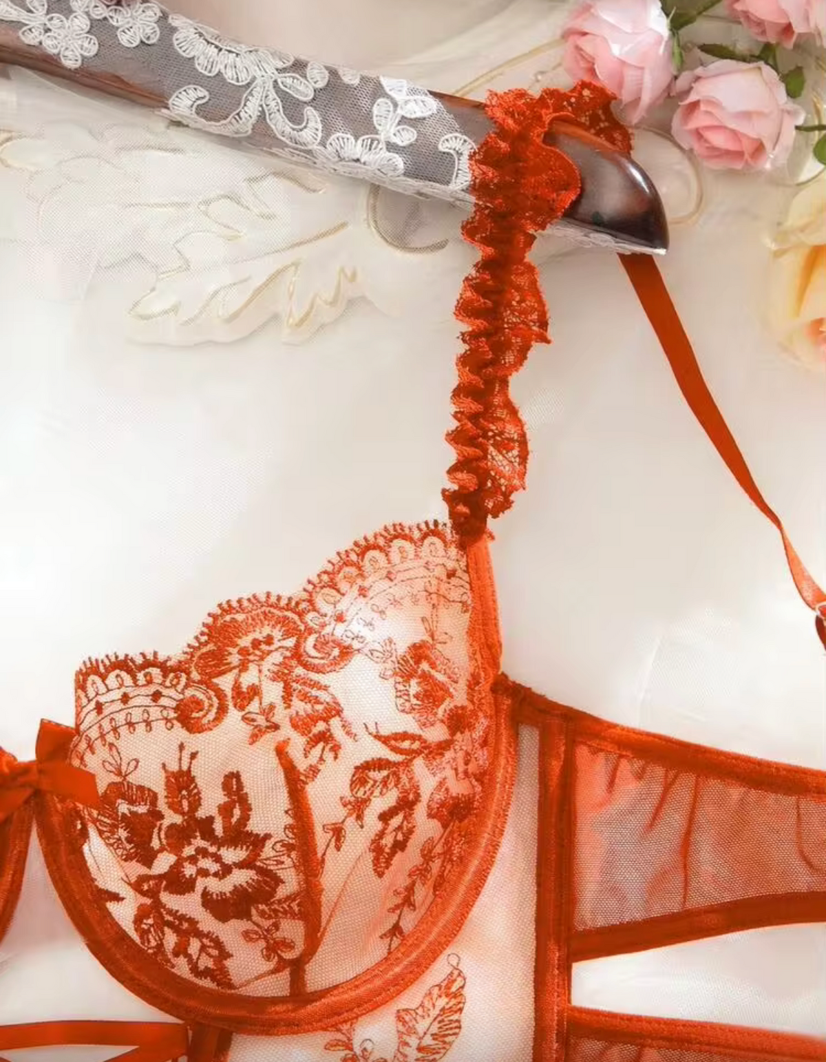 An orange lace bralette with sheer mesh and ruffled straps, shown in a close-up product photo with decorative flowers and a patterned ribbon