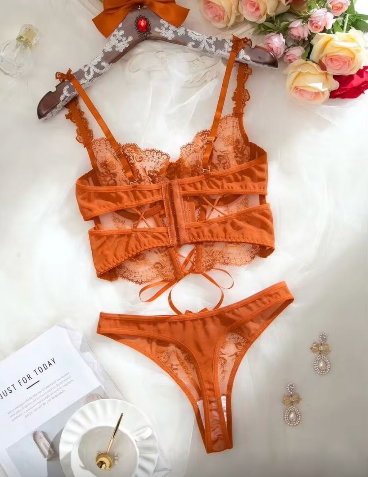 An orange lace two-piece lingerie set consisting of a strappy bralette and matching panties, displayed on a white fabric background with decorative flowers, a teacup, and a hanger.