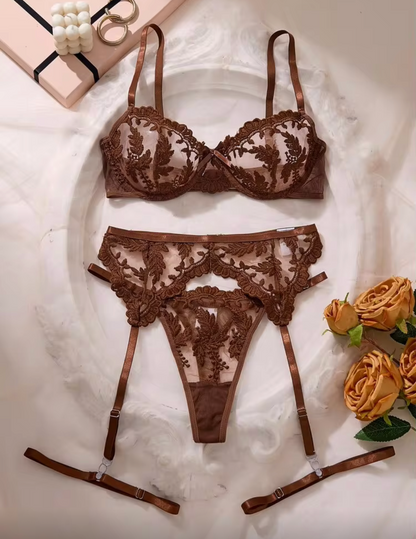 A three-piece brown lace lingerie set laid flat on a white fabric background, featuring a bra, matching panties, and garter straps. Decorative gold rings and yellow roses are placed around the set.